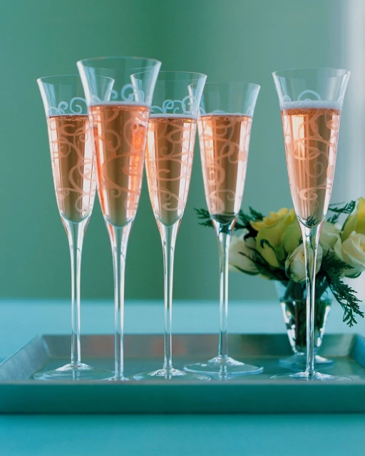 Elegant flute glasses with decorative swirls filled with rose champagne on a reflective surface, accented by white flowers, soft lighting.