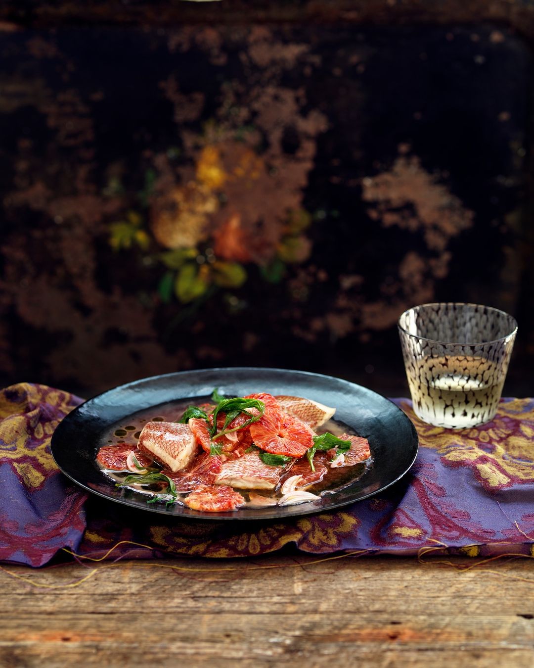 Overhead shot of a black plate featuring a grapefruit salad with fresh herbs, accompanied by a spotted glass on a wooden table.