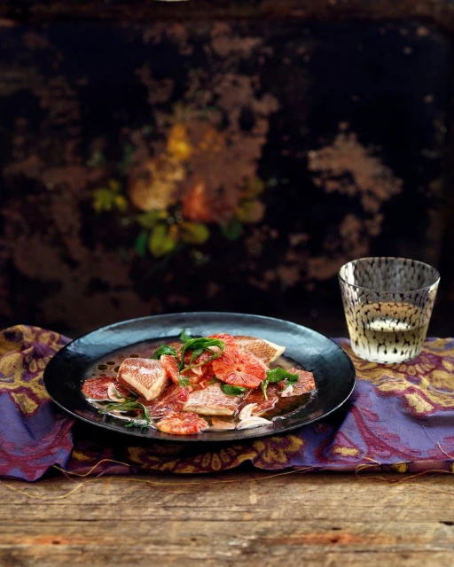 Overhead shot of a black plate featuring a grapefruit salad with fresh herbs, accompanied by a spotted glass on a wooden table.