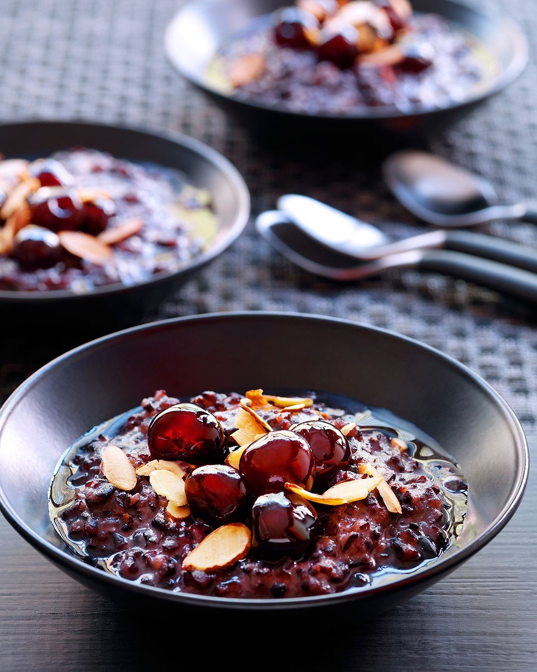 Three bowls of black rice pudding are garnished with cherries and sliced almonds, and spoons rest on a woven grey surface.