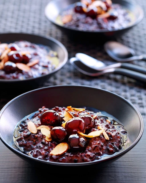 Three bowls of black rice pudding are garnished with cherries and sliced almonds, and spoons rest on a woven grey surface.