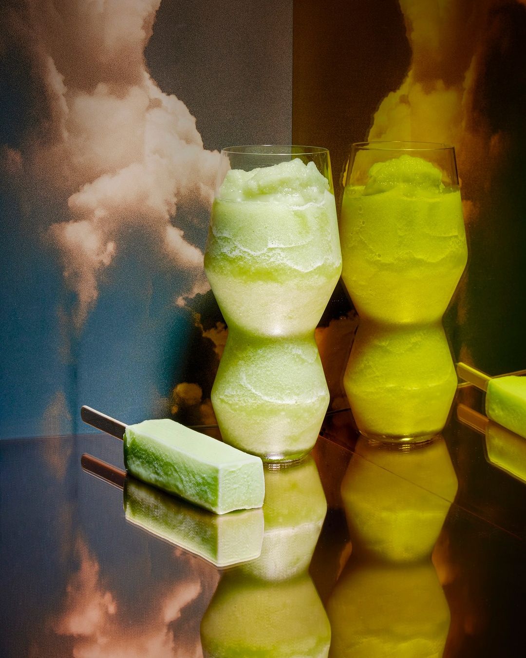 Two glasses filled with green and yellow slushie drinks, with matching popsicles, set against an abstract sky backdrop on reflective surface.