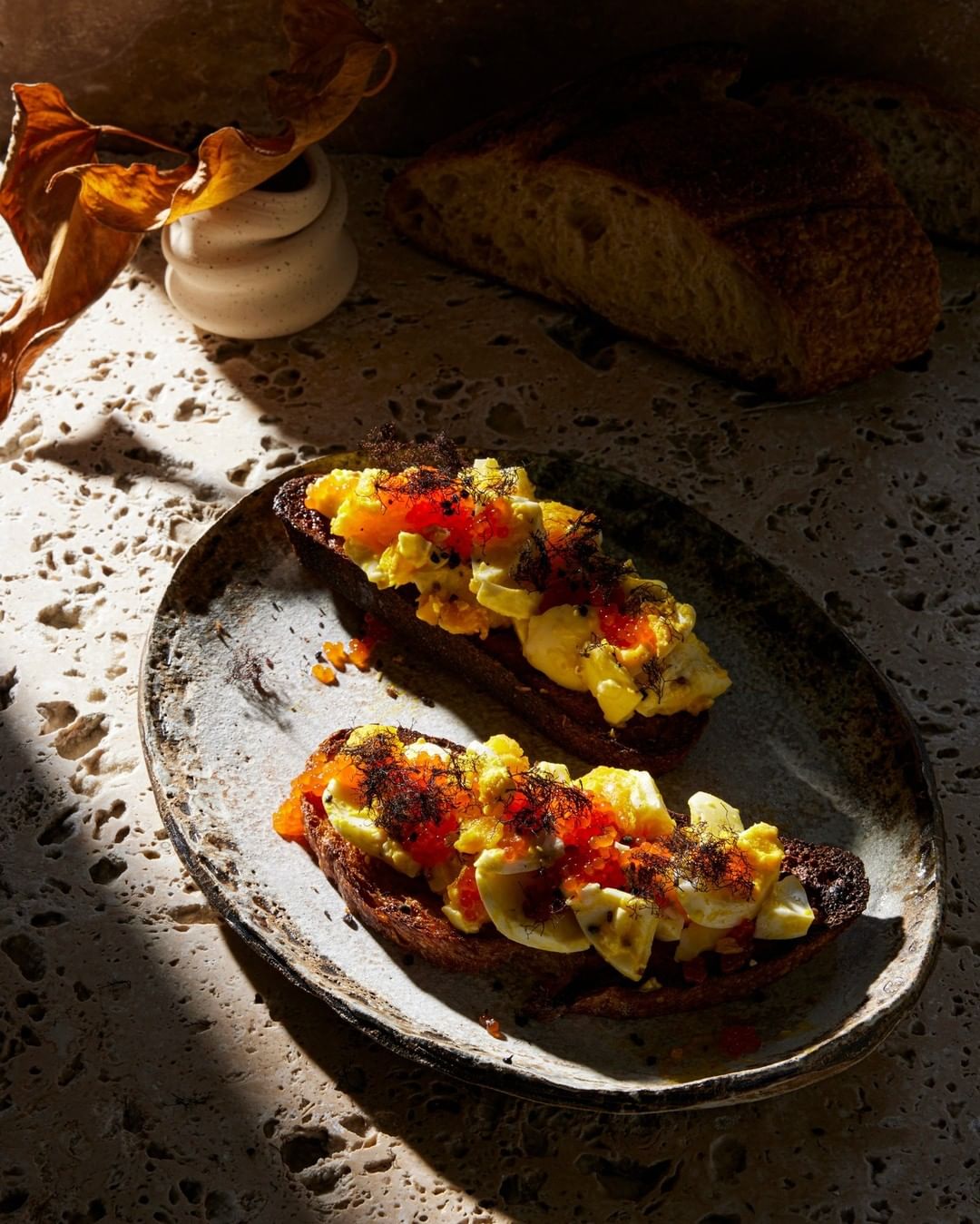 Two open-faced toasts with scrambled eggs and orange fish roe, presented on a ceramic plate with a neutral-toned textural backdrop.