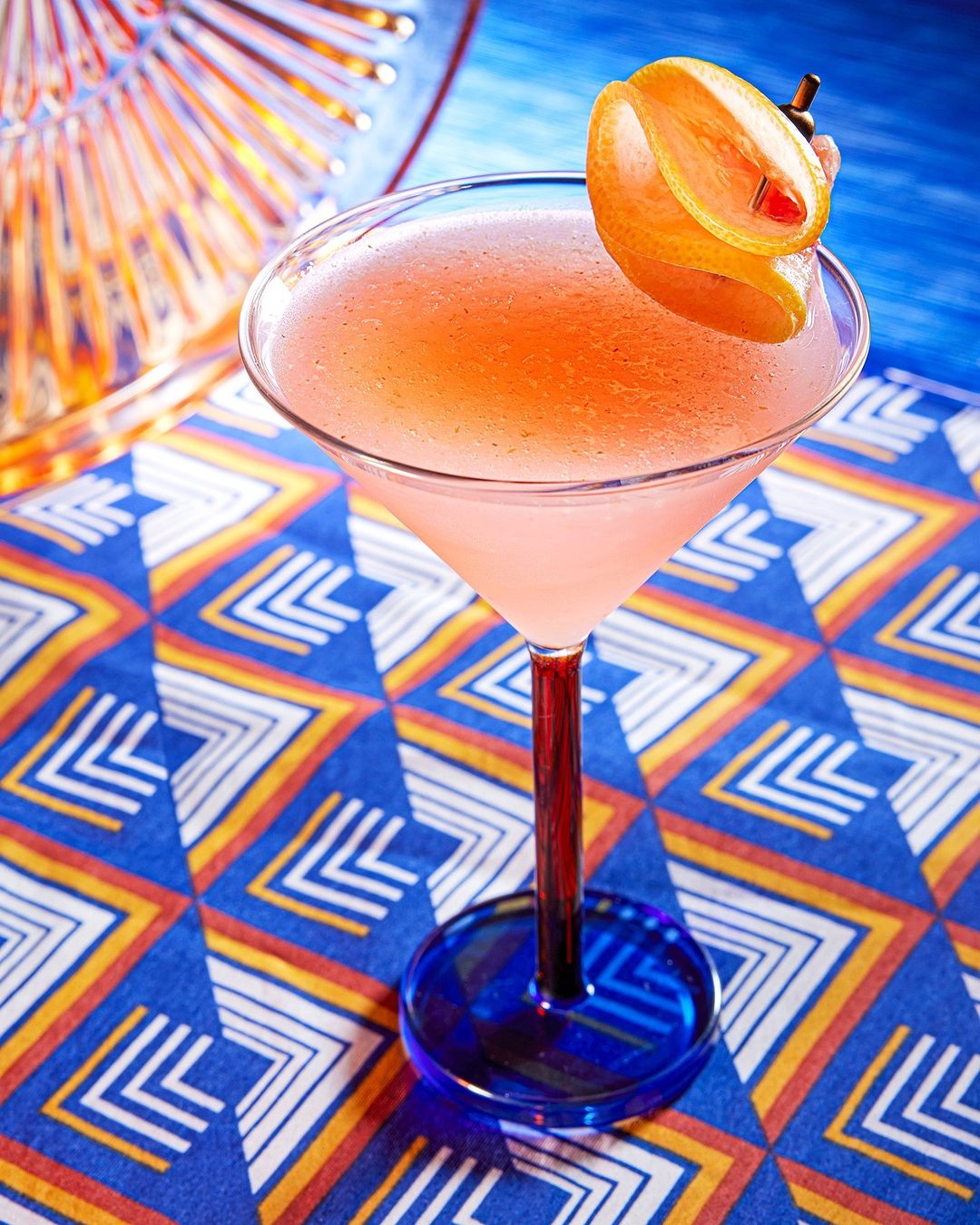 Close-up of a vibrant pink cocktail in a stemmed glass, garnished with a grape curl, set against a patterned blue cloth backdrop.