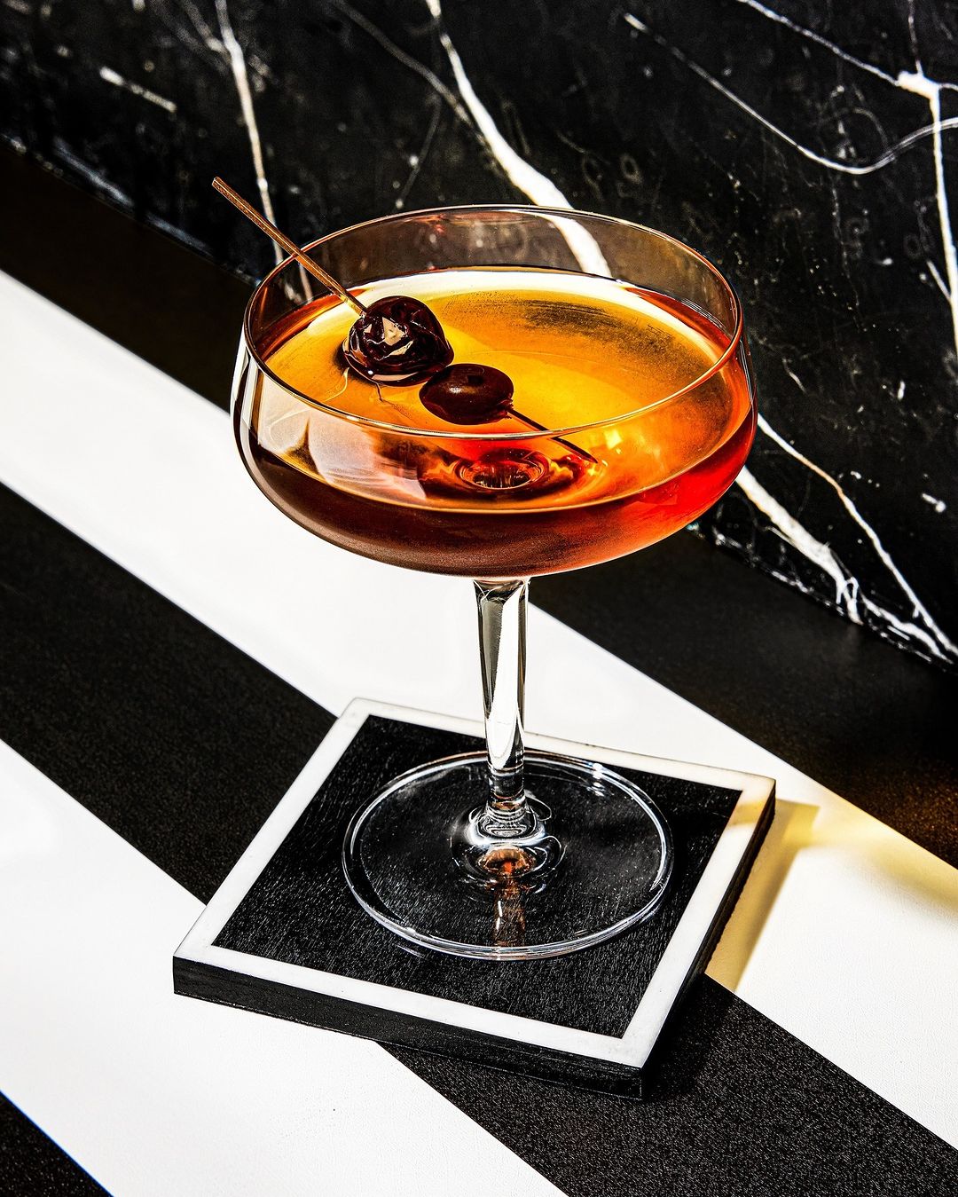 Stemmed cocktail glass on a black and white striped surface. The glass contains an amber-colored cocktail with two cherries.