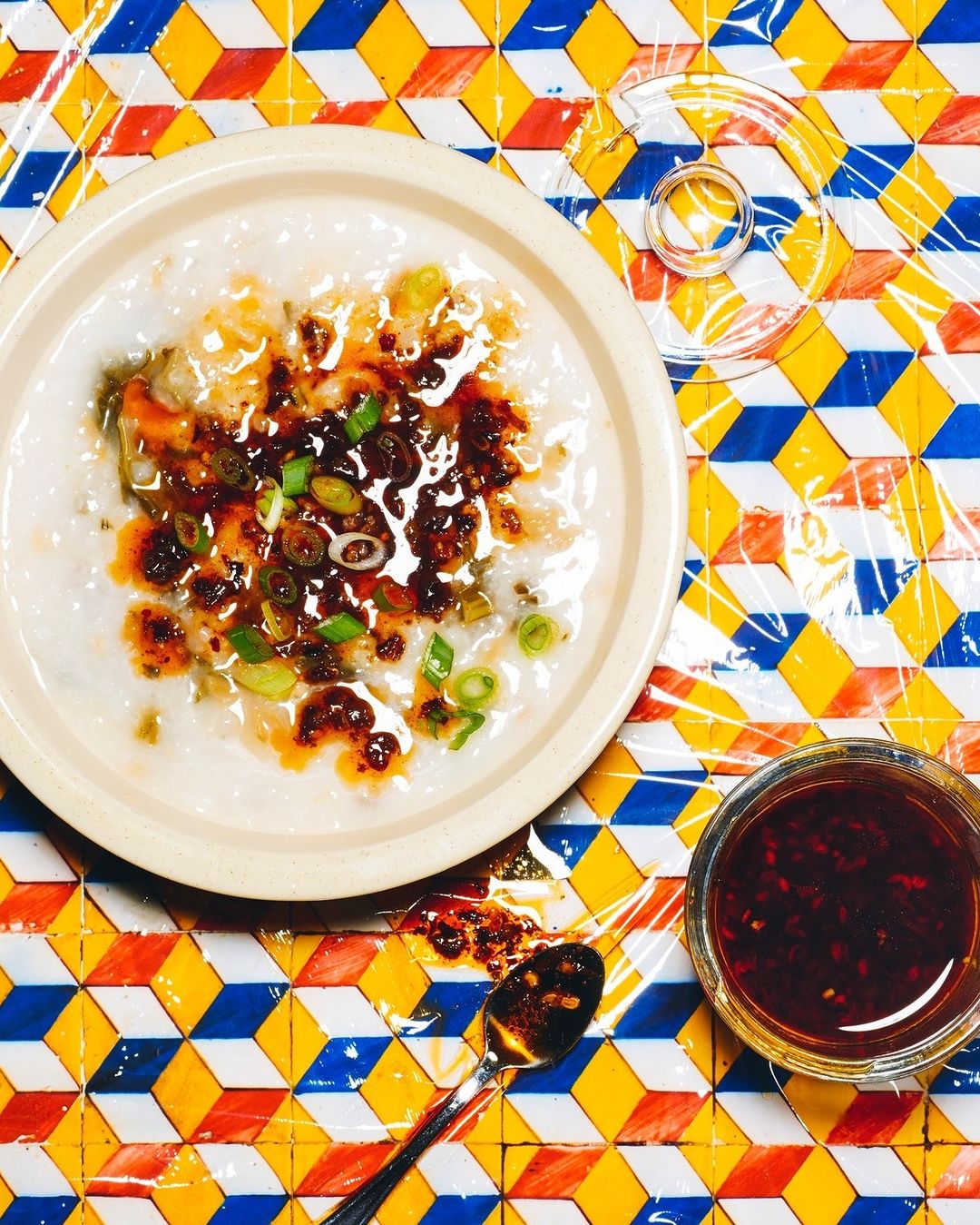 Overhead shot of a bowl of congee topped with chili oil and green onions on a colorful, geometric table with a spoon and chili oil jar.