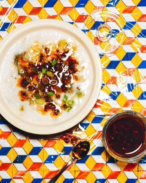 Overhead shot of a bowl of congee topped with chili oil and green onions on a colorful, geometric table with a spoon and chili oil jar.