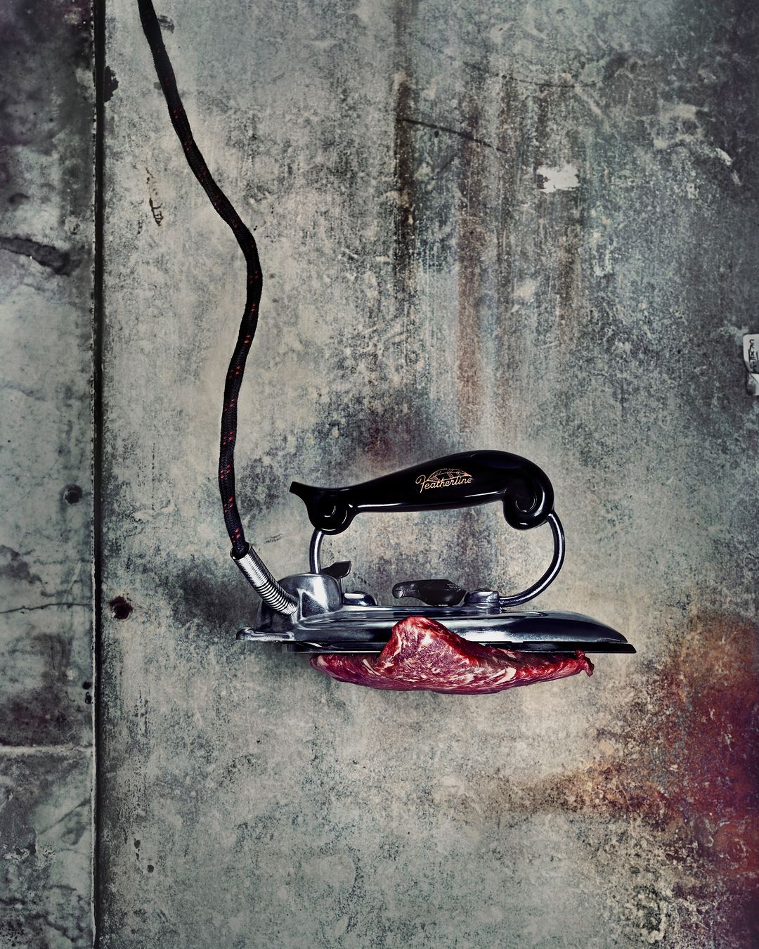 Close-up of vintage electric iron pressing raw meat against a textured grey wall, creating a surreal and unconventional image.