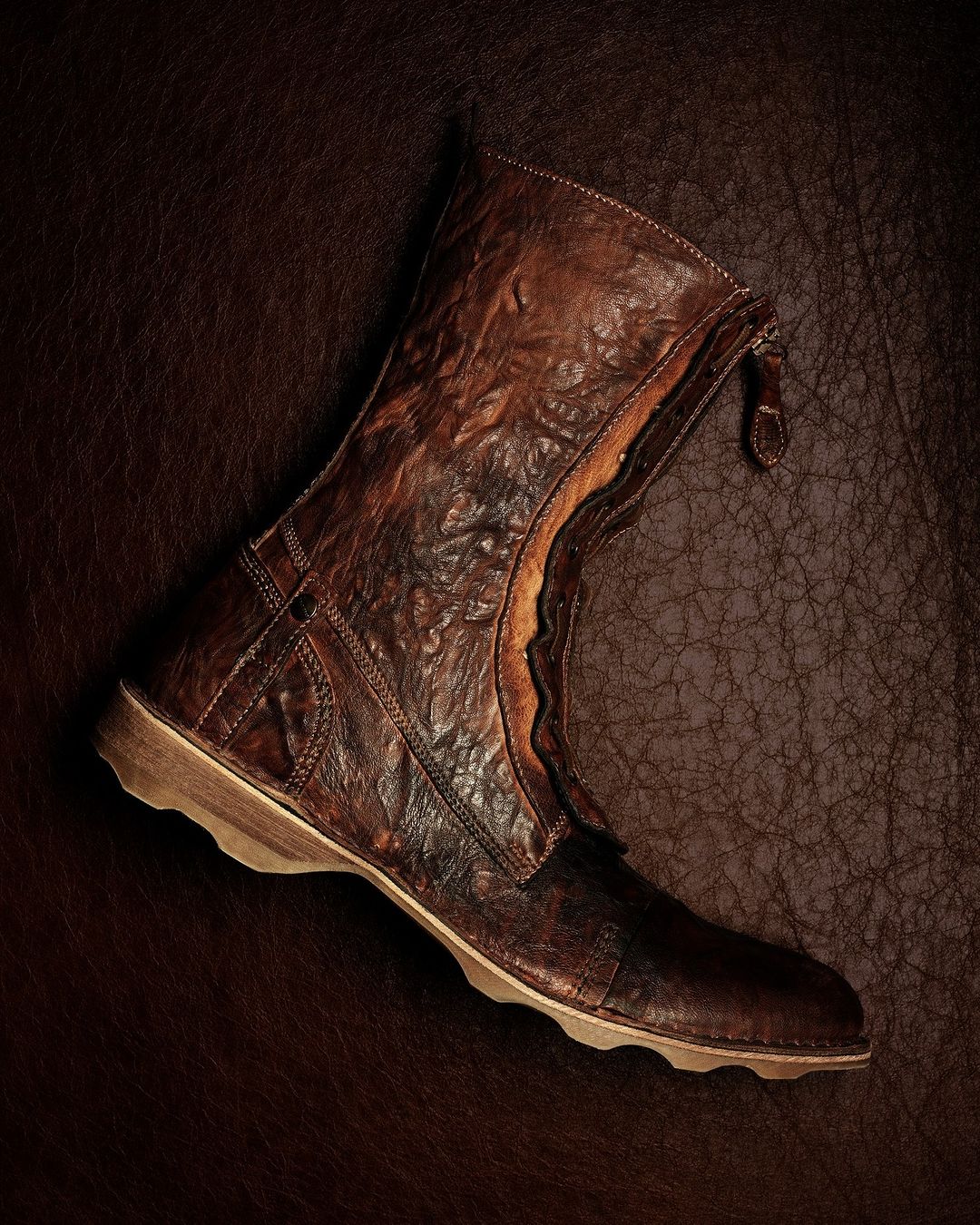 Detailed product shot of a distressed brown leather ankle boot with a zipper closure on a matching textured leather backdrop.