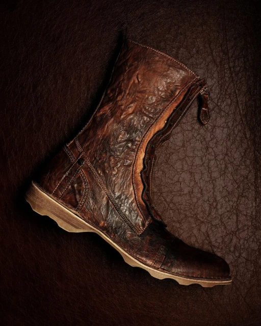 Detailed product shot of a distressed brown leather ankle boot with a zipper closure on a matching textured leather backdrop.