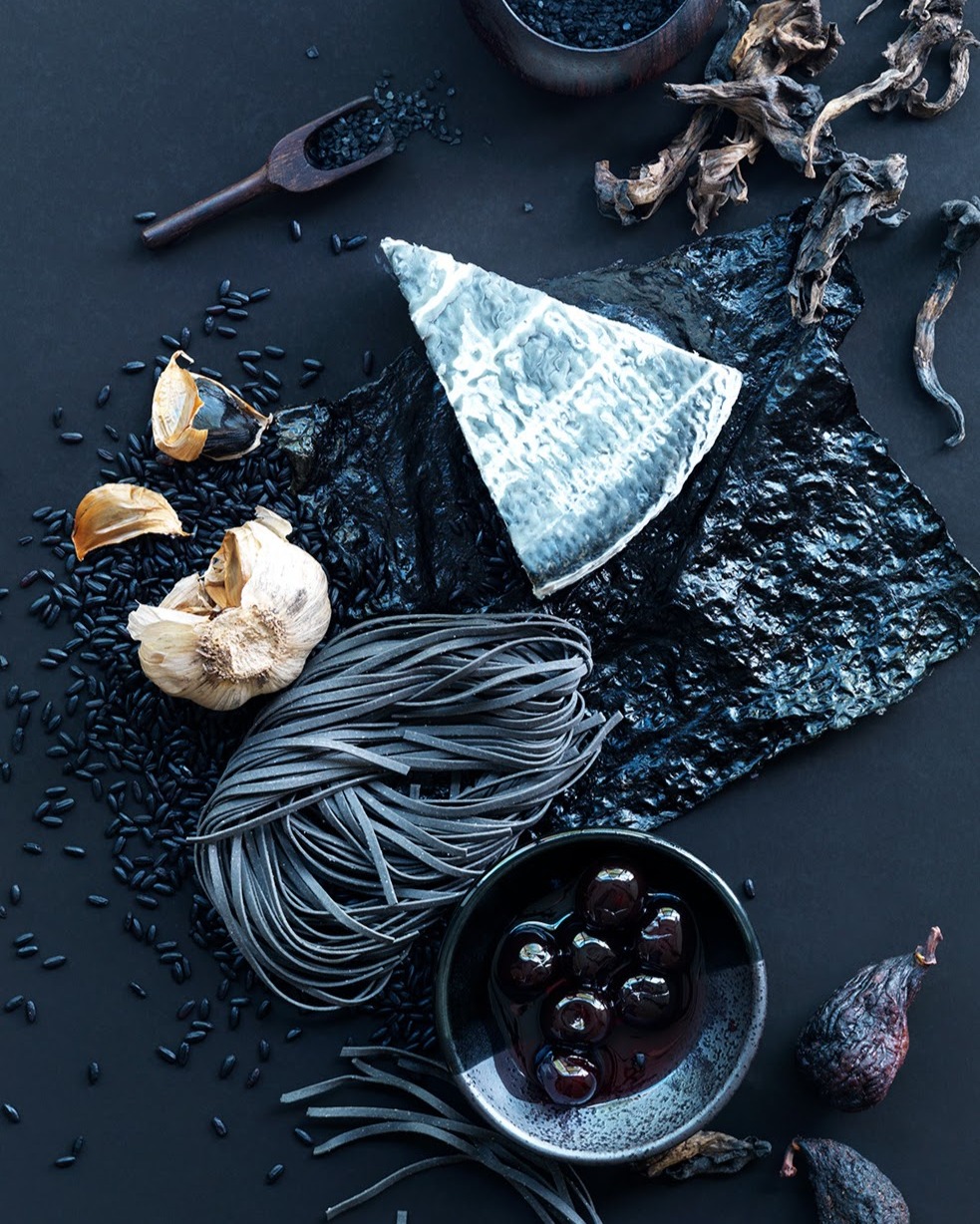 Still life food photography featuring black linguine pasta, a wedge of white cheese, black garlic, and black rice on a dark surface.