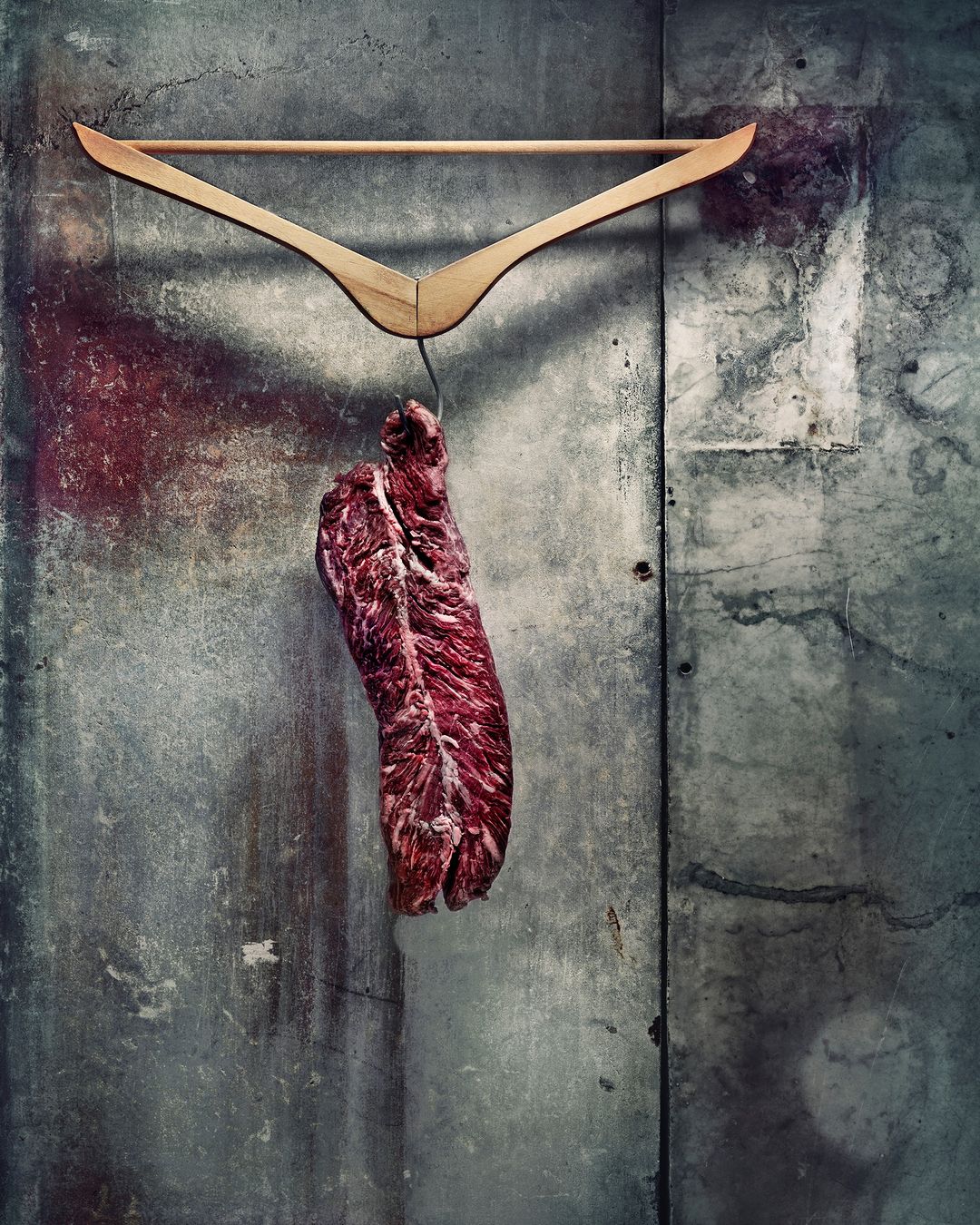 Raw meat hangs from a wooden hanger against a textured concrete wall, showcasing rustic food presentation and simple visual composition.