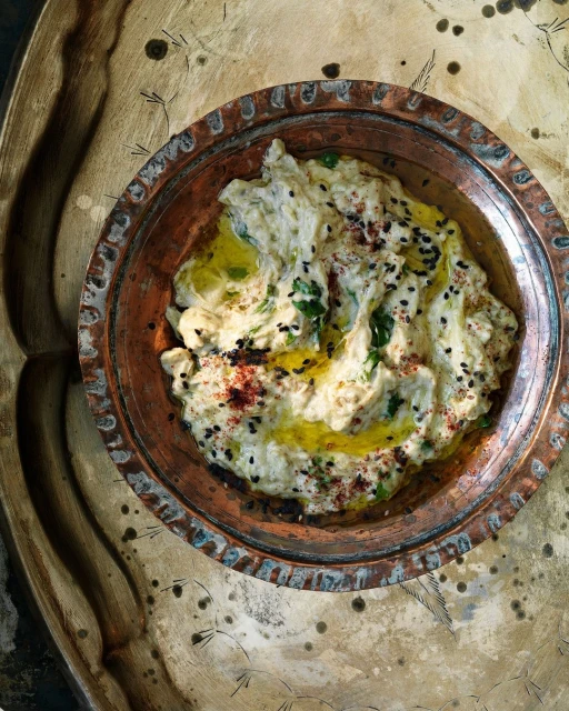 Overhead shot of creamy baba ghanoush in a copper bowl, drizzled with olive oil, herbs, black sesame seeds, and red pepper flakes.