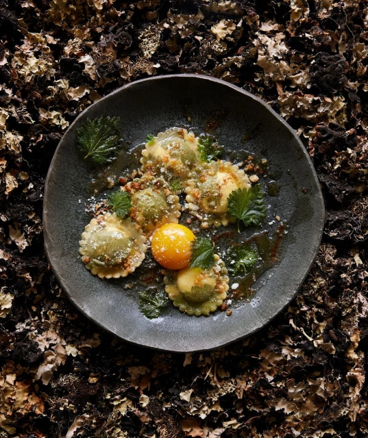 Overhead shot of green ravioli with an egg yolk on a dark plate, set against a textured dark brown and tan organic background.