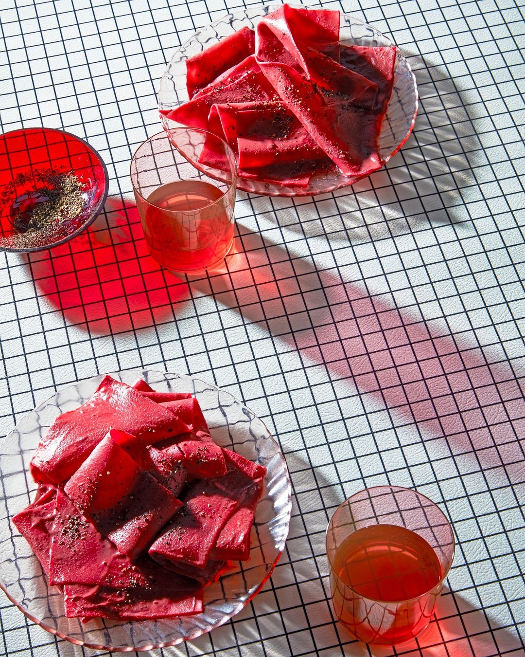Two glass plates of red fruit leather and clear drinking glasses are positioned on a black and white checkered tablecloth.
