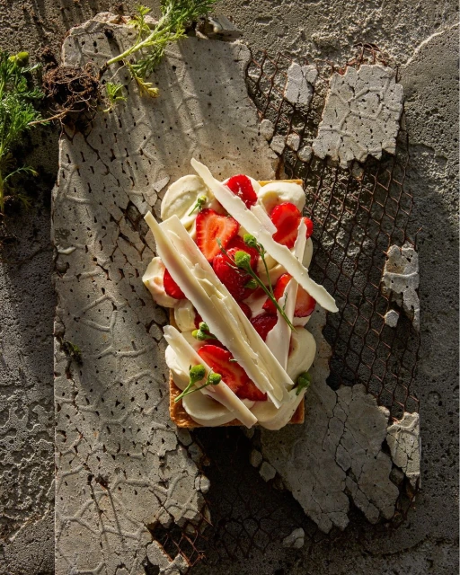 Overhead shot of a strawberry tart with white chocolate shavings and cream on a textured gray surface with foliage.