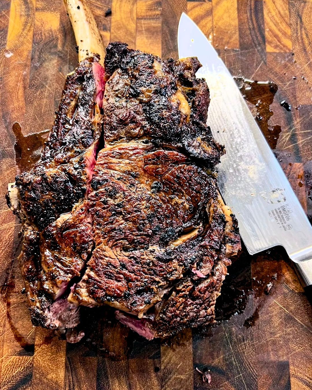 Close-up of a grilled tomahawk steak on a wooden cutting board with a large knife; rich texture and dark char marks visible.