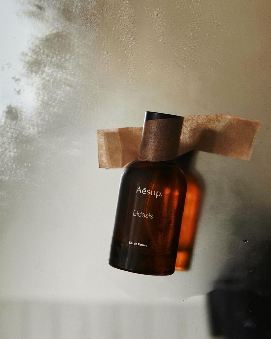 A dark amber glass bottle of Aesop Eidesis Eau de Parfum is displayed against a softly blurred background and held in place with tape.