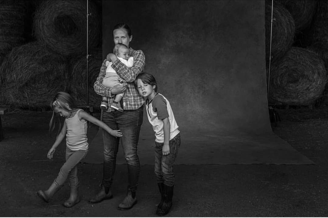 Black and white studio shot of a woman holding a baby, with two children standing nearby in an indoor setting with hay bale background.