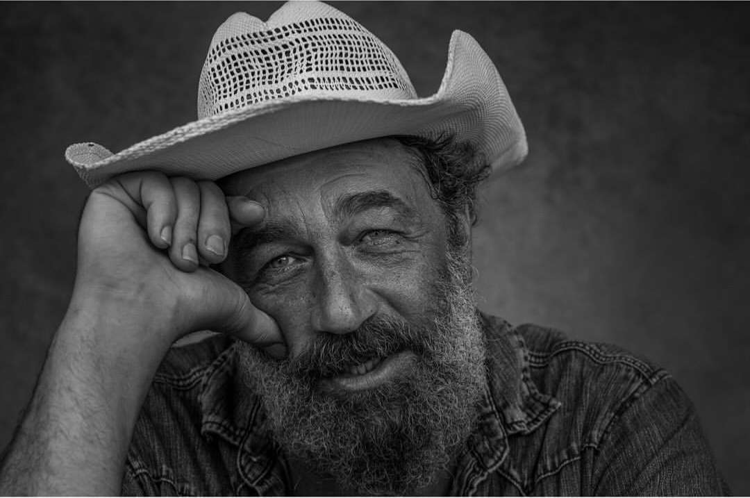 Portrait of an adult male with a beard wearing a cowboy hat, with a soft smile, captured in a black-and-white photo studio setting.