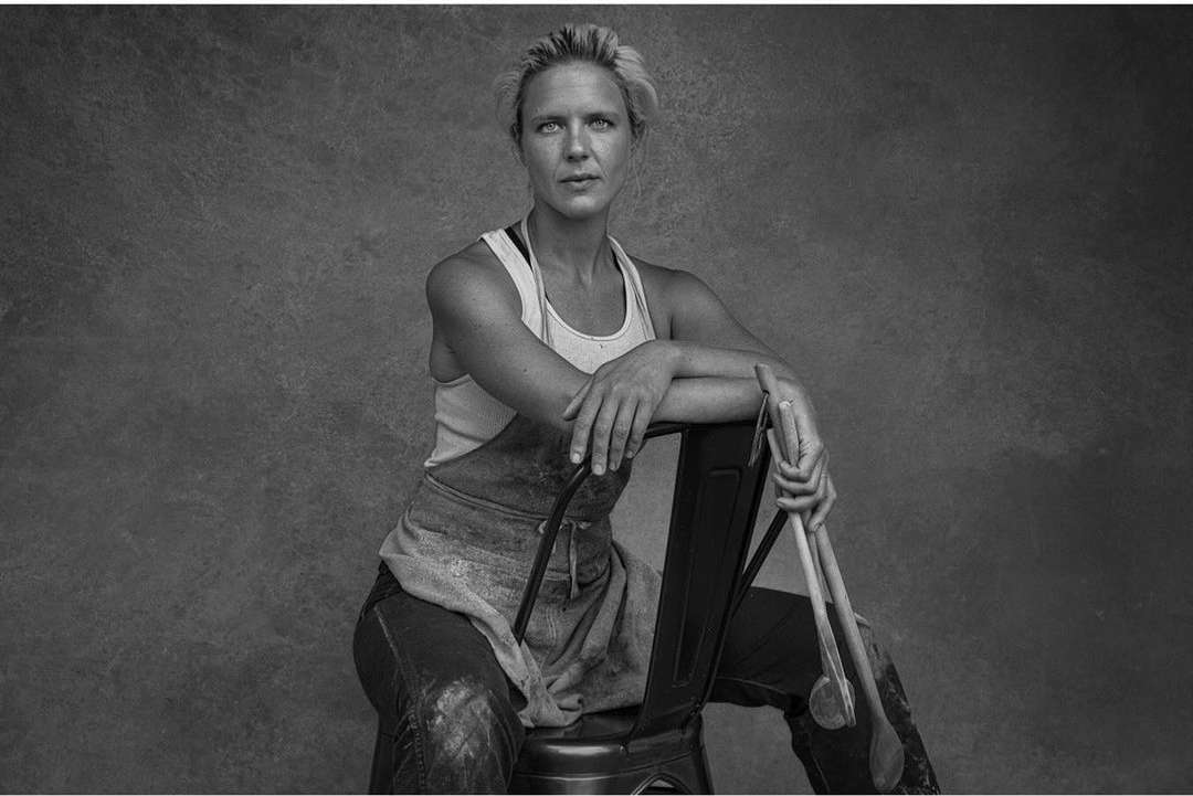 Monochrome portrait of a fair-skinned adult female wearing an apron and holding wooden spoons, sitting on a metal chair against a textured wall.