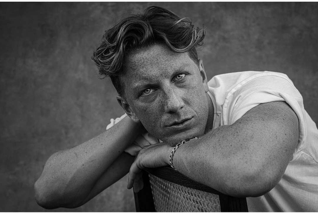 Black and white portrait of an adult male with curly hair leaning on a chair, looking directly at the camera against a dark background.