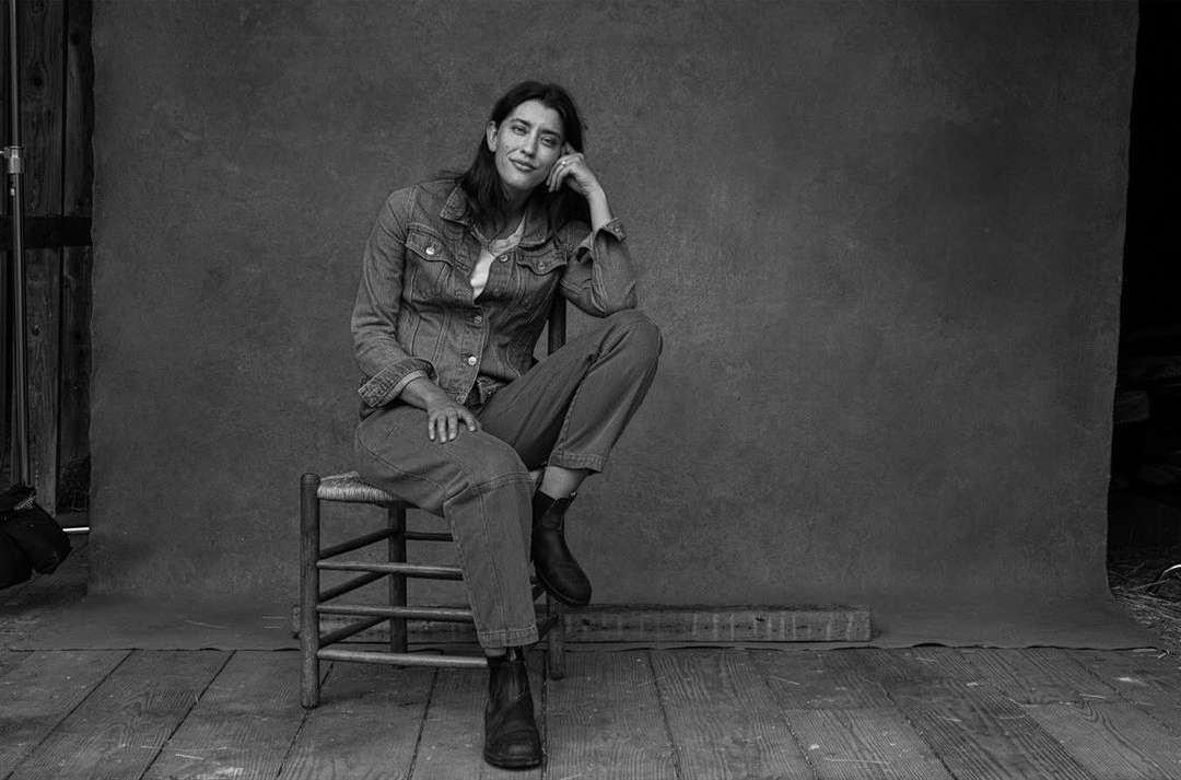 Black and white portrait of a woman in a denim jacket and jeans sitting on a wooden chair in front of a textured gray backdrop.