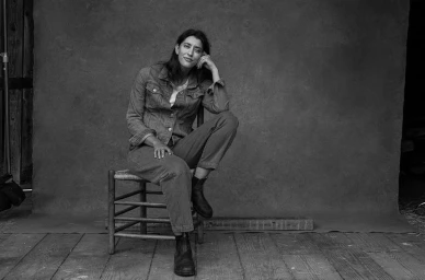 Monochrome portrait of a woman seated on a wooden chair wearing a denim jacket and pants against a textured backdrop