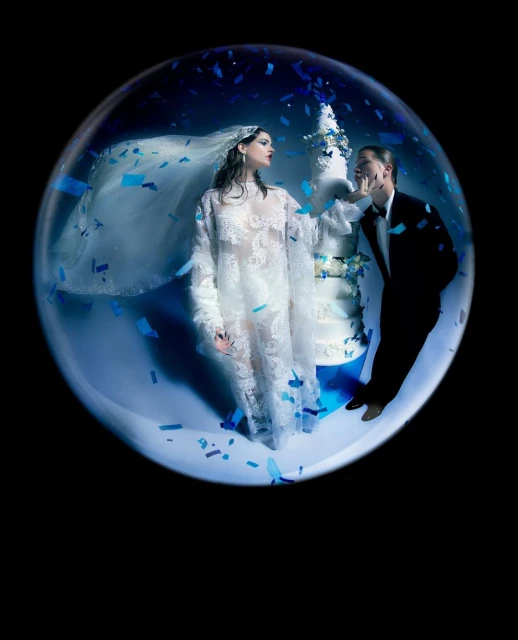 A couple stands inside a large clear bubble with confetti around them, the woman in white lace and the man in a black tuxedo.