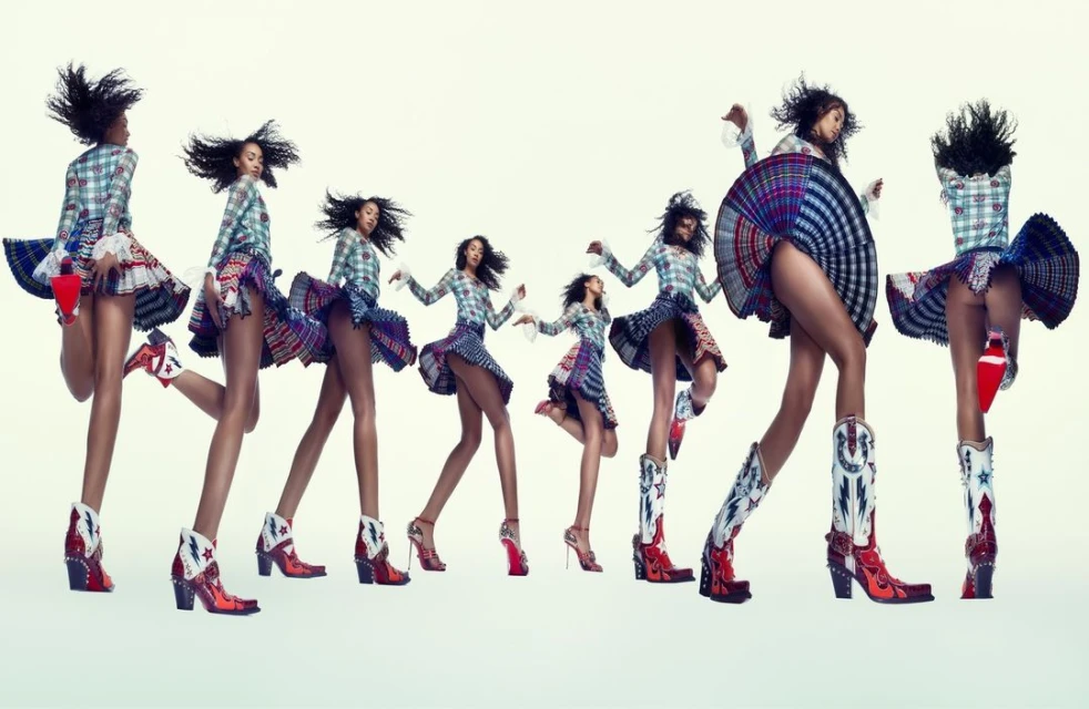 A diverse group of women in plaid tops and short skirts perform a choreographed dance against a pale backdrop, showcasing their legs and boots.