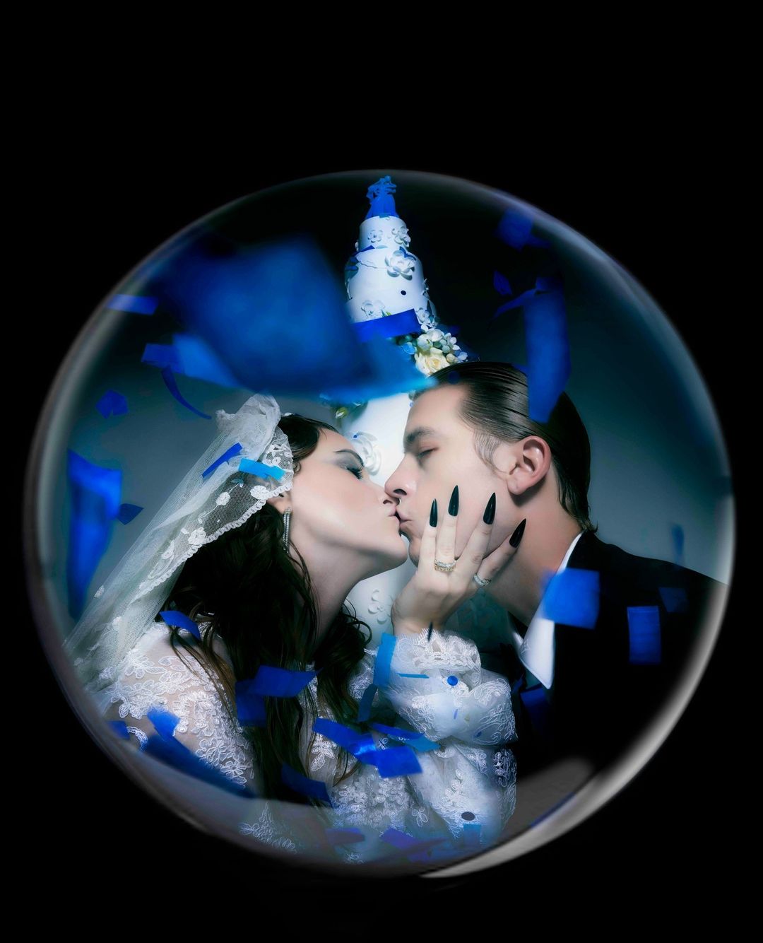 A bride and groom kissing inside a clear bubble with a tiered wedding cake in the background, surrounded by blue confetti.
