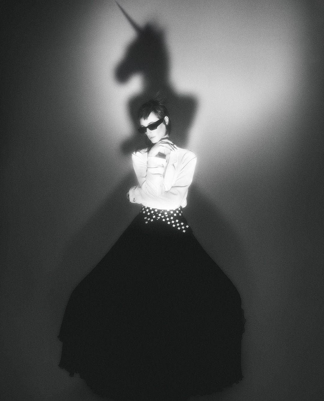 Monochrome studio portrait of a woman in sunglasses, white top, and long black skirt, with a unicorn shadow projected on the background.
