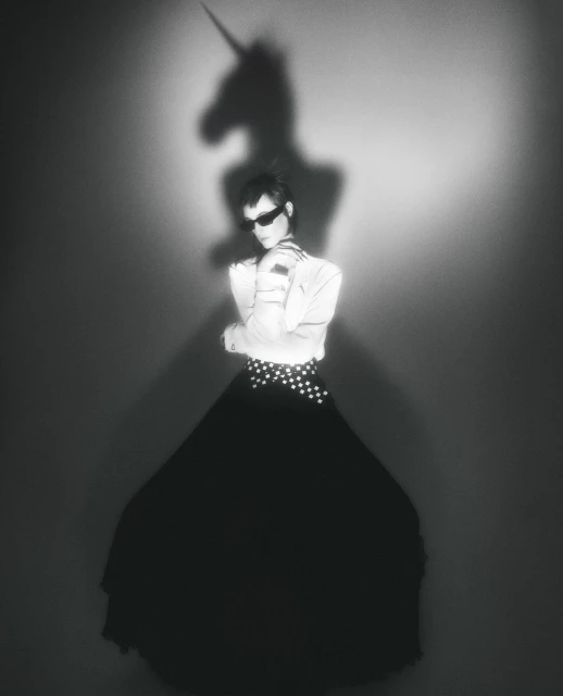 Monochrome studio portrait of a woman in sunglasses, white top, and long black skirt, with a unicorn shadow projected on the background.