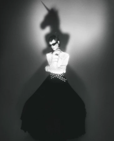 Monochrome studio portrait of a woman in sunglasses, white top, and long black skirt, with a unicorn shadow projected on the background.