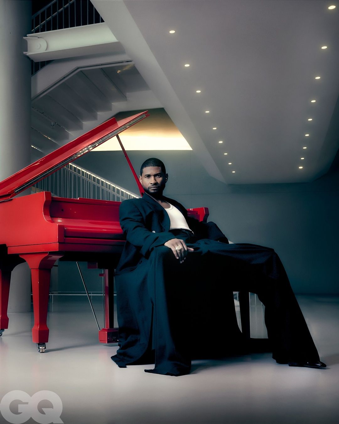 Portrait of Usher sitting next to a red grand piano, wearing a black suit in a modern architectural setting with a staircase and lighting.