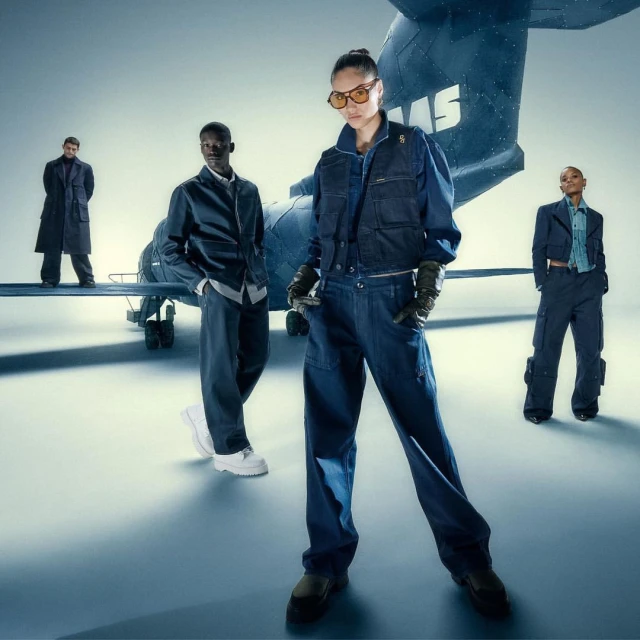 Four fashion models in stylish denim and navy blue outfits pose in front of a small aircraft in a brightly lit studio setting.
