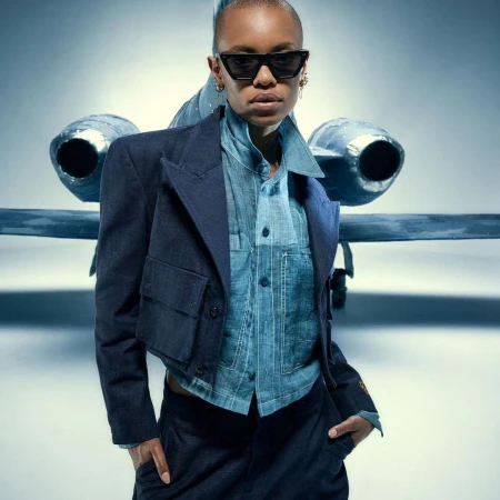 Stylish woman poses in front of airplane wearing a navy blazer, denim shirt, dark pants, sunglasses, and jewelry in a studio.