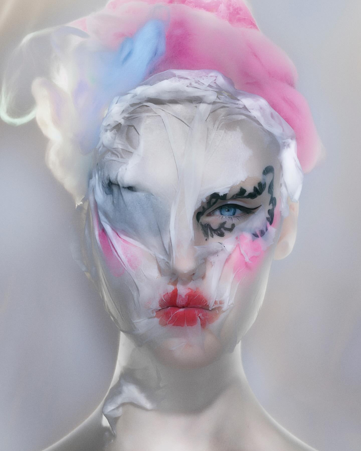 Artistic image of a woman with elaborate makeup, translucent plastic wrap, and a pastel floral headpiece against a soft grey background.