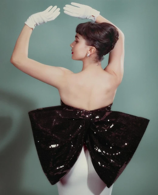Woman in a strapless white dress with a black sequin bow, wearing white gloves, posing against a teal background in a studio setting.