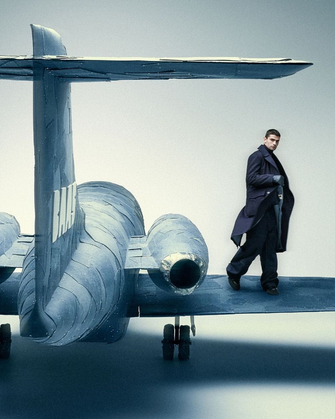 An adult male stands on the wing of a blue airplane against a gradient white backdrop, wearing a navy trench coat and dark trousers.