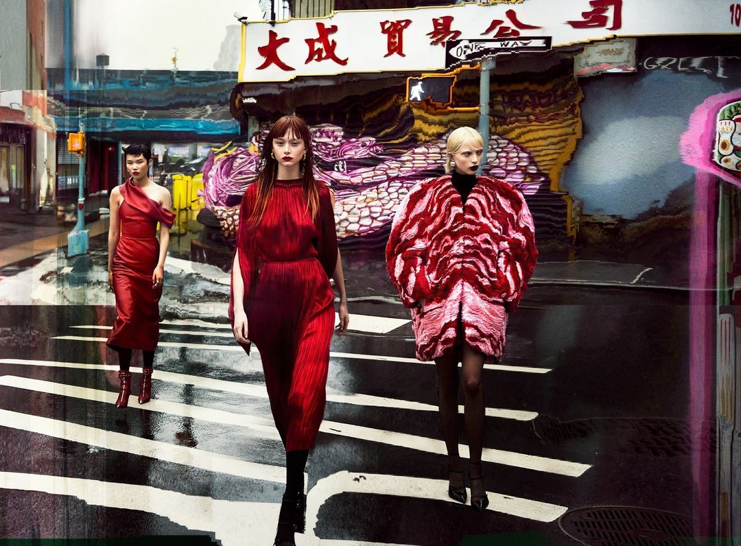 Three fashion models in red dresses pose on a city street with a crosswalk and a building featuring East Asian text in the background.