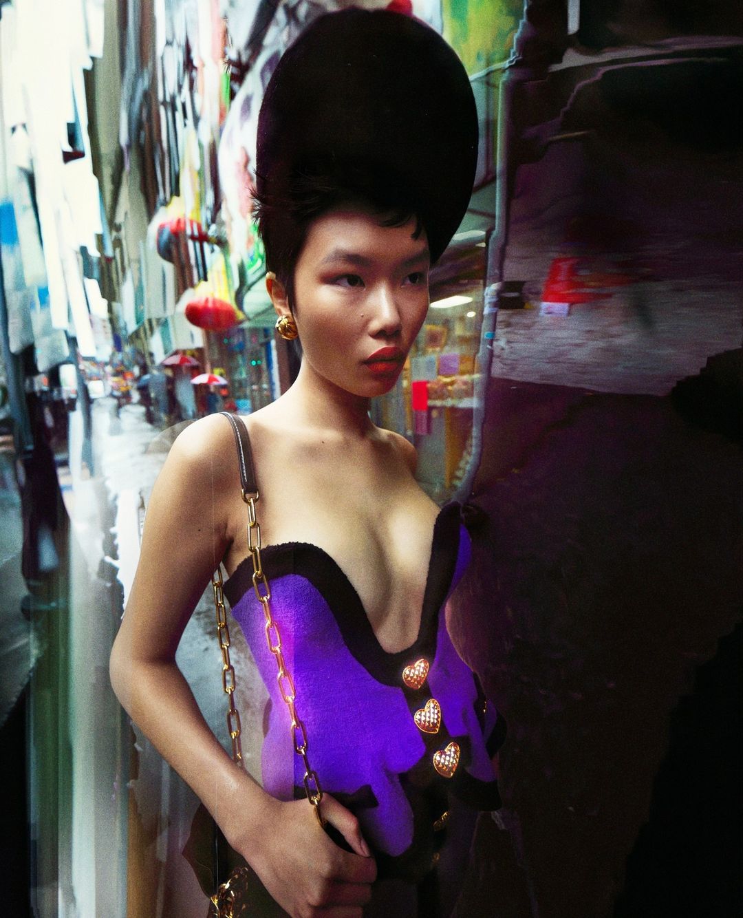 Asian woman in purple corset and black hat poses on city street with blurred buildings and umbrellas in the background for a fashion shoot.
