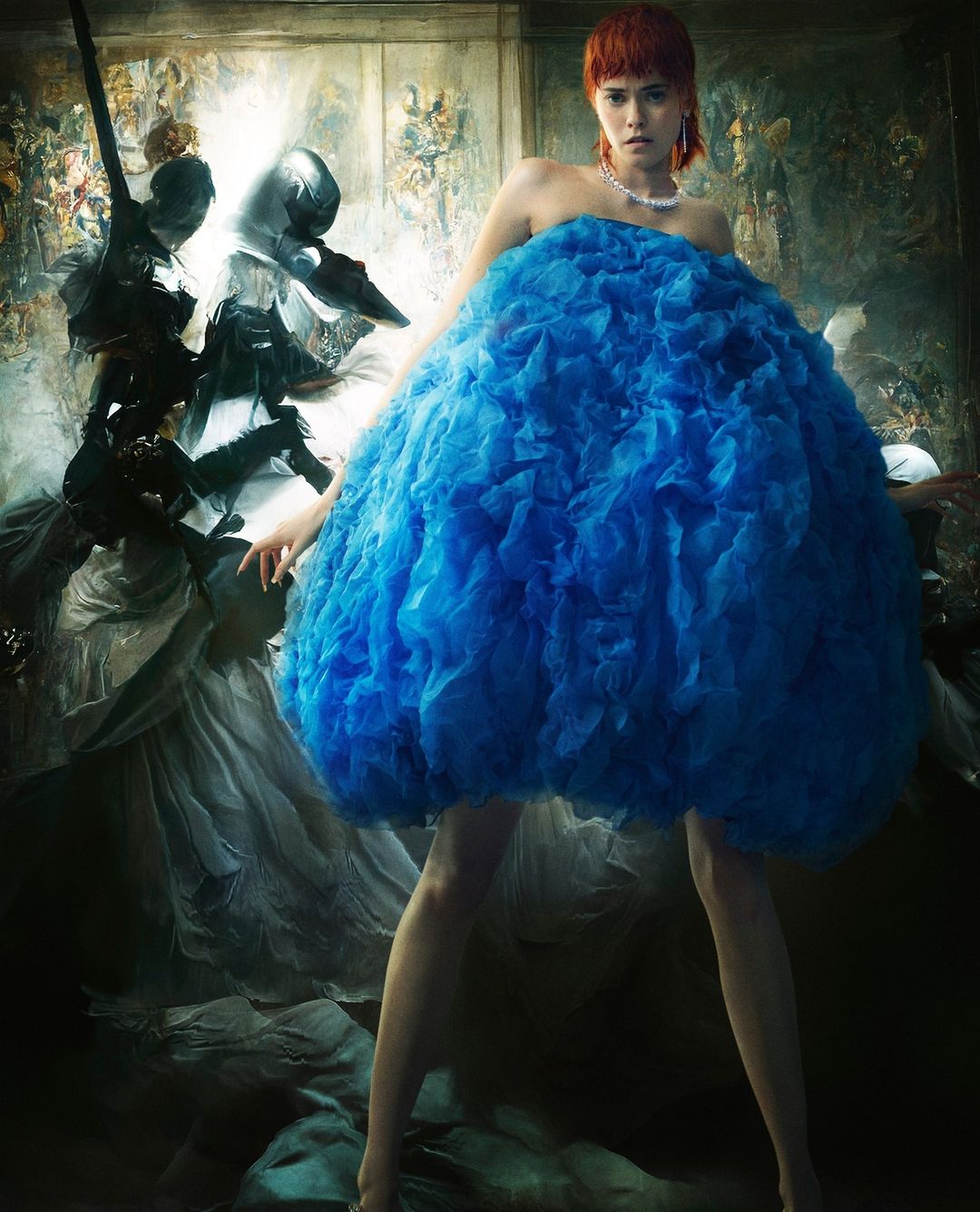 Fashion shot of a red-haired adult female wearing an elaborate, frilly, bright blue dress in a vintage studio setting with a muted palette.