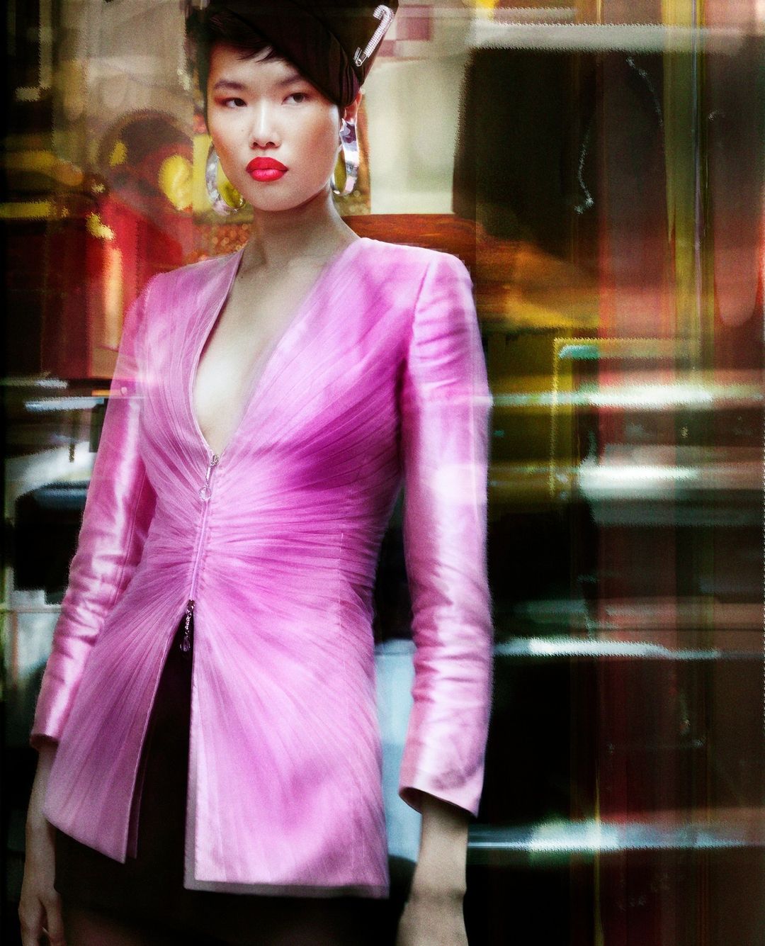 Model in a bright pink structured jacket with large silver hoop earrings and vibrant red lipstick against a blurred background