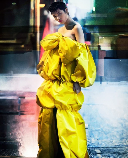 Fashionable person in a yellow oversized dress, posing in an abstract urban setting with light reflections and textures, creating a striking image.