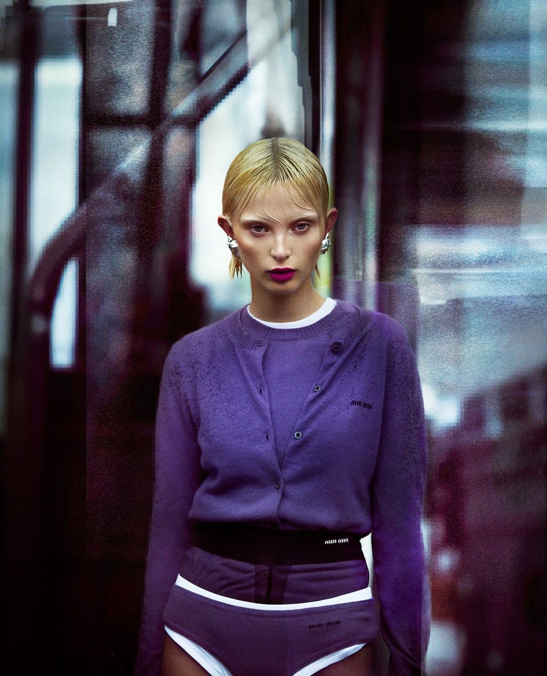 A blonde female fashion model in a purple cardigan and shorts with white trim poses in front of a blurry background, fashion shot.