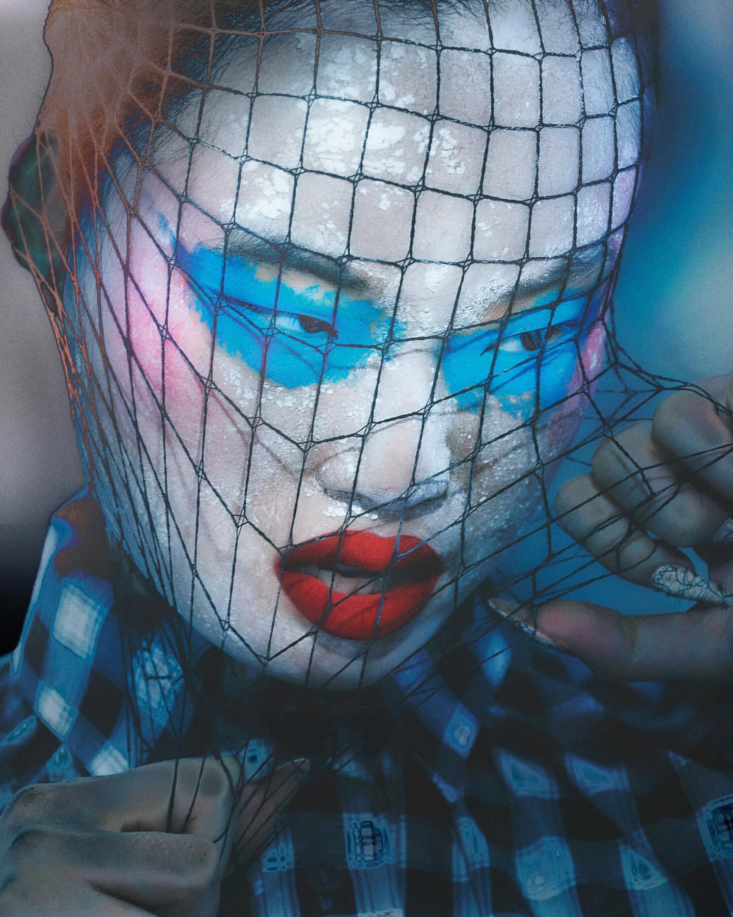 Person with blue and pink face paint under a black net, wearing a blue plaid shirt with red lipstick in a studio setting.