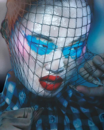 Person with blue and pink face paint under a black net, wearing a blue plaid shirt with red lipstick in a studio setting.