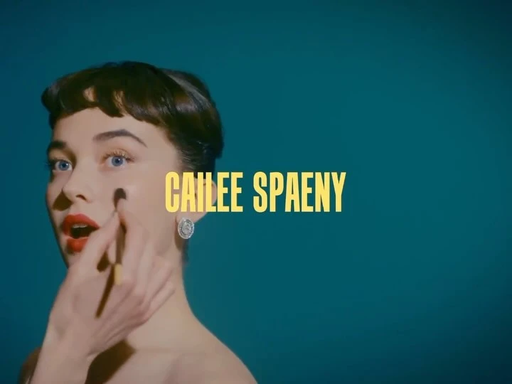Glamorous young woman applying makeup with red lipstick and styled hair against teal backdrop with the name Cailee Spaeny.