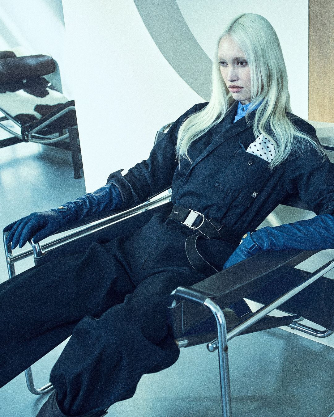 A young adult female with platinum blond hair poses in a dark blue utility suit and leather chair inside a photo studio with graphic background.